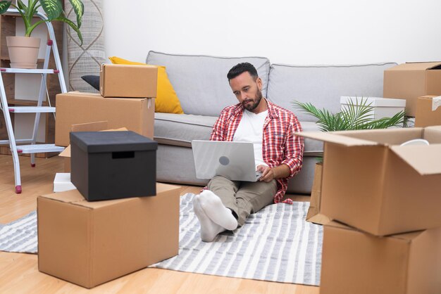 Hombre se sienta en medio de cajas portátil en la mano de compras en línea para decorar el apartamento en movimiento