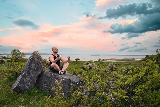 un hombre se sienta en una roca frente a un lago y mira al mar.