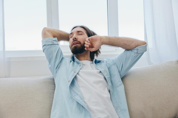 Un hombre se sienta en el sofá bostezando y estirándose después de una siesta falta de sueño y fatiga del trabajo y rutina diaria inadecuada foto de alta calidad