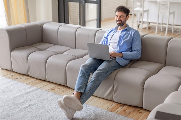 Un hombre se sienta en un sofá modular gris en una sala de estar moderna usando una computadora portátil que está sonriendo y