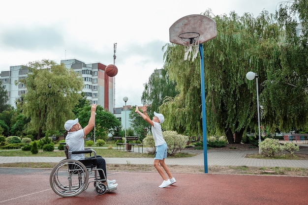 Un hombre en silla de ruedas juega baloncesto con su hijo en el campo de deportes. Padre discapacitado, infancia feliz, concepto de persona discapacitada.