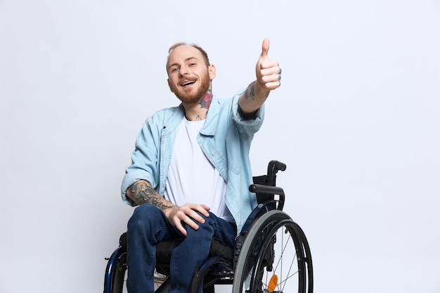 Un hombre en silla de ruedas mira a la cámara y muestra un pulgar hacia arriba de felicidad con tatuajes en las manos sentado en un estudio gris concepto de salud hombre con discapacidad