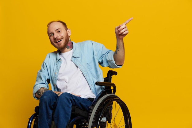 Foto un hombre en silla de ruedas con problemas con el sistema musculoesquelético mira a la cámara y muestra un dedo con tatuajes en las manos sentado en un fondo de estudio amarillo concepto de salud hombre con discapacidad