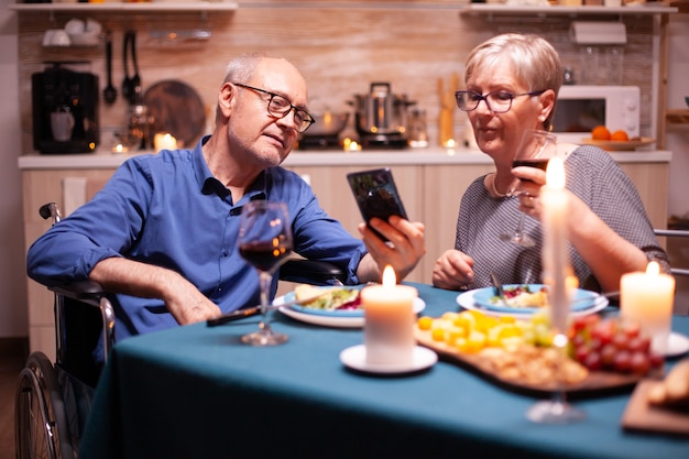 Hombre en silla de ruedas sosteniendo el teléfono mientras cena con su esposa en la cocina. Desplazamiento y visualización de fotos. Marido senior discapacitado inmovilizado regañando por teléfono disfrutando de la comida festiva.