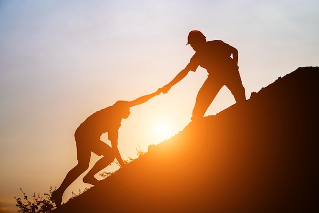 Hombre silueta ayudando a un amigo a escalar una montaña contra el cielo durante la puesta de sol