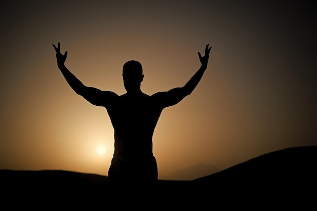 Foto hombre con silueta en forma en el cielo del atardecer. deportista con mano levantada sobre paisaje natural. el atleta muestra el poder. entrenamiento en el atardecer de verano. concepto de deporte, bienestar y cuidado corporal.