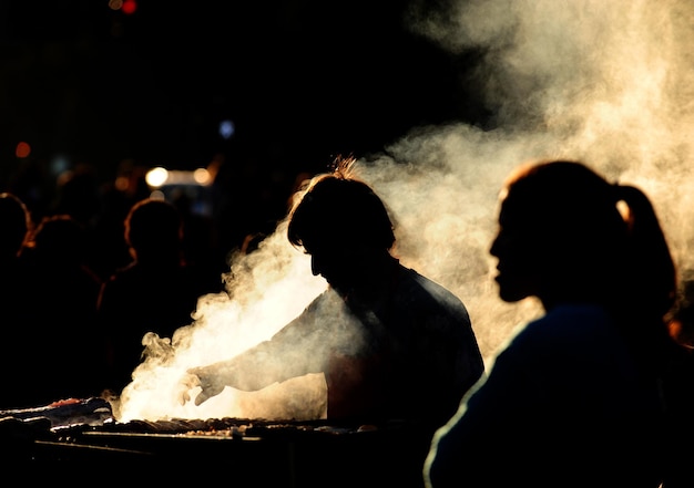 Hombre silueta de pie con la gente mientras prepara comida en la parrilla de barbacoa en medio del humo
