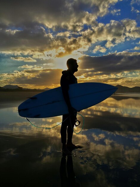 Foto hombre silueta de pie con la tabla de surf en la orilla en la playa contra el cielo nublado durante la puesta de sol