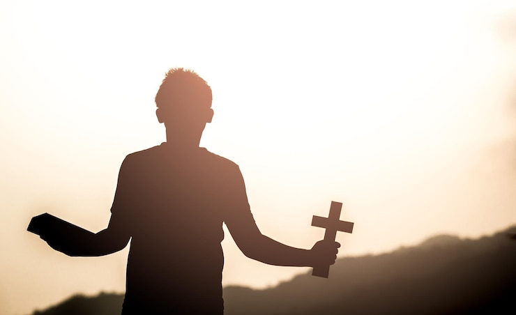 Foto hombre silueta sosteniendo un crucifijo y una biblia contra el cielo durante la puesta del sol