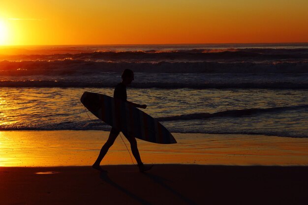 Foto hombre silueta con tabla de surf caminando por la orilla en la playa contra el cielo durante la puesta de sol