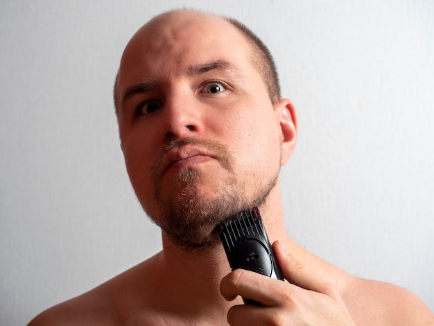 Un hombre sobre un fondo gris se afeita la barba con una maquinilla de afeitar eléctrica. Mira a la cámara, recortándose el pelo. Belleza y cuidado masculino en casa. luz dura