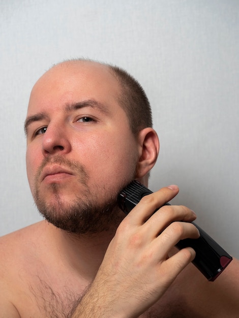 Un hombre sobre un fondo gris se afeita la barba con una maquinilla de afeitar eléctrica.