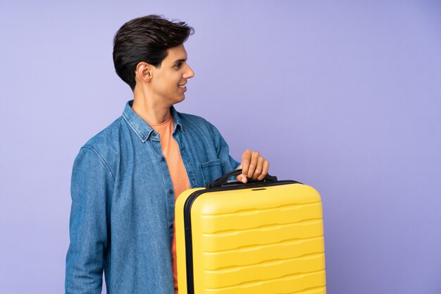 Hombre sobre pared púrpura aislada en vacaciones con maleta de viaje y un sombrero