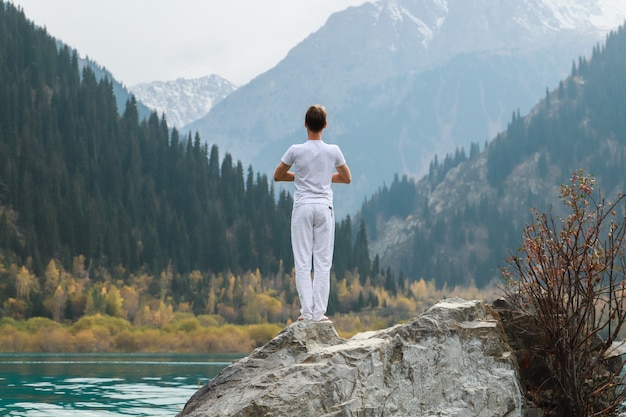 Un hombre se para sobre una piedra en el centro de un lago de montaña y practica yoga. Pose Vrikshasana.