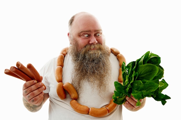 Un hombre con sobrepeso con un montón de salchichas alrededor del cuello mirando las espinacas y sosteniendo las salchichas por otro lado, intenta elegir lo que es más saludable.