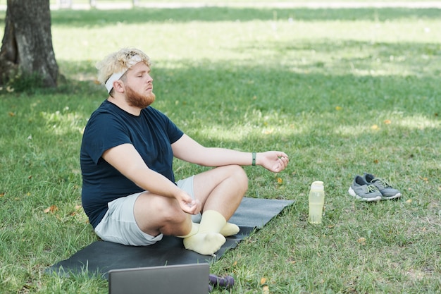Hombre con sobrepeso sentado en la colchoneta con los ojos cerrados y hacer yoga en el parque