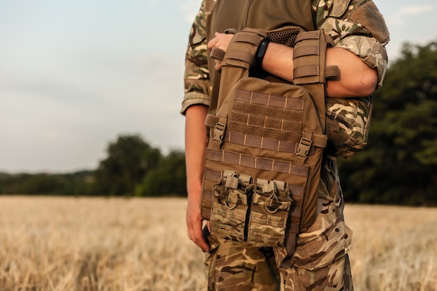 Hombre soldado de pie contra un campo. Soldado en traje militar con chaleco antibalas. Foto de un soldado en traje militar sosteniendo una pistola y chaleco antibalas sobre fondo naranja del desierto.