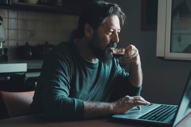 Un hombre solo en casa usando la computadora por la noche en la cocina y bebiendo café para no quedarse dormido Insomnio y atención saludable Hombres que trabajan demasiado con una computadora portátil y un trabajo moderno en línea Estrés en línea