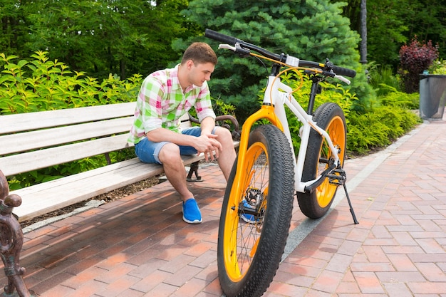 Hombre soltero solitario en pantalones cortos con expresión triste sentado en un banco cerca de la bicicleta con neumáticos gordos estacionados en la acera de ladrillo en el hermoso parque verde