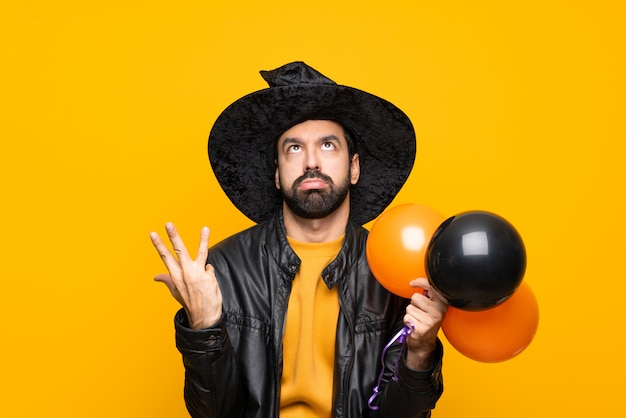 Hombre con sombrero de bruja sosteniendo globos de aire negros y naranjas para la fiesta de halloween frustrado por una mala situación