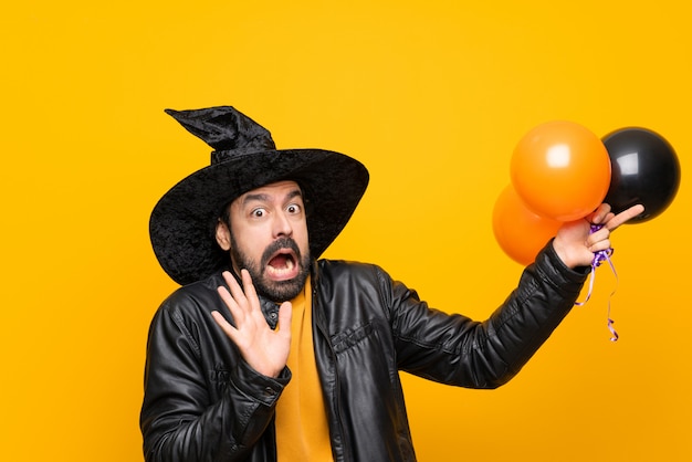 Hombre con sombrero de bruja sosteniendo globos de aire negros y naranjas para la fiesta de halloween nervioso y asustado