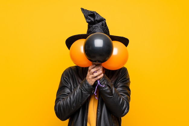 Hombre con sombrero de bruja sosteniendo globos negros y naranjas para la fiesta de halloween cubriendo los ojos con las manos