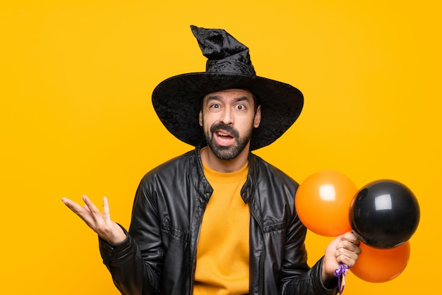 Hombre con sombrero de bruja sosteniendo globos negros y naranjas para fiesta de halloween haciendo dudas gesto