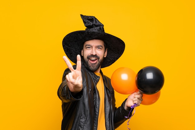 Hombre con sombrero de bruja sosteniendo globos negros y naranjas para la fiesta de halloween sonriendo y mostrando el signo de la victoria