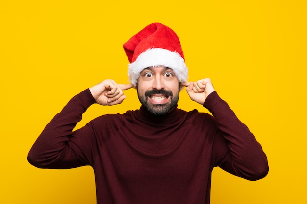 Hombre con sombrero de Navidad sobre pared amarilla aislada frustrado y cubriendo las orejas