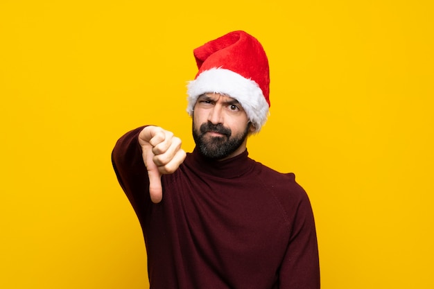 Hombre con sombrero de Navidad sobre pared amarilla aislada mostrando el pulgar hacia abajo con expresión negativa