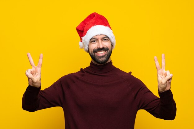 Hombre con sombrero de Navidad sobre pared amarilla aislada que muestra el signo de la victoria con ambas manos