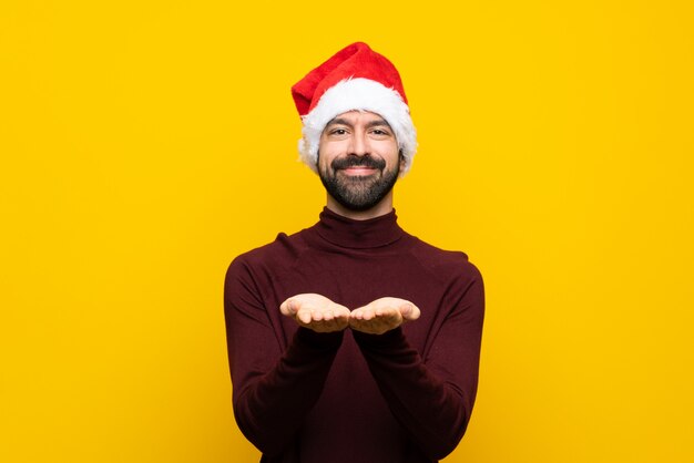 Hombre con sombrero de Navidad sobre pared amarilla aislada sosteniendo copyspace imaginario en la palma para insertar un anuncio