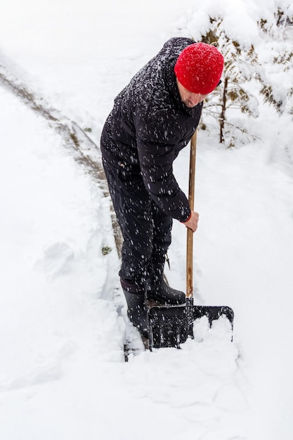 Un hombre con un sombrero rojo limpia el camino de la nieve con una pala en un día de invierno en una nevada