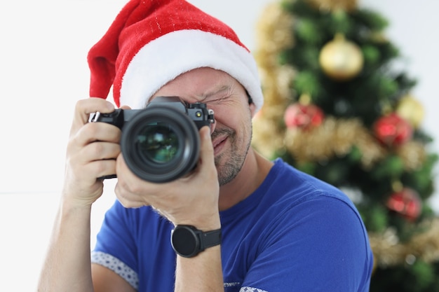 Hombre con sombrero rojo de santa tomando fotografías con la cámara cerca del árbol de navidad