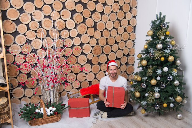 Hombre con sombrero de santa con sonrisa de caja roja en el piso. Árbol de Navidad, trineo, canasta, nieve, arbusto de frutos rojos, decoraciones. navidad, año nuevo, obsequios y obsequios. Feliz dia del boxeo. Concepto de celebración de vacaciones.