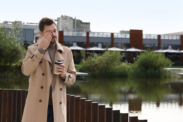 Hombre soñoliento con una taza de café cerca del río al aire libre Espacio para texto