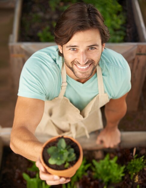 Foto hombre sonríe y planta desde arriba para apreciar el regalo o la venta en la tienda desde un ángulo alto jardiner masculino feliz y sosteniendo la vegetación en la olla para el crecimiento de decoración o el cultivo en la guardería