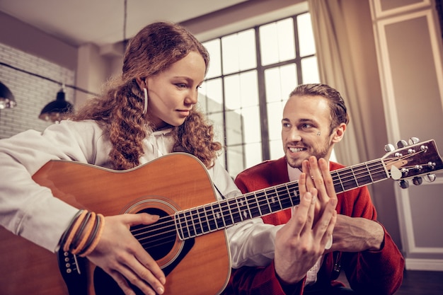 Hombre sonriendo. Hombre de pelo oscuro con barba sonriendo mientras escucha a su alumno tocando la guitarra