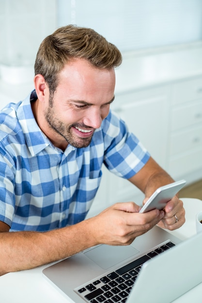 Hombre sonriendo mientras usa el teléfono celular por computadora portátil en la mesa en la cocina