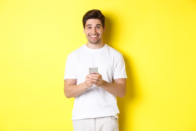 Hombre sonriendo y mirando feliz después de leer la oferta promocional en el teléfono inteligente, de pie contra la pared amarilla en camiseta blanca.