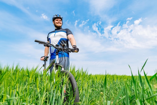 Hombre sonriente adulto joven de pie con bicicleta en cebada verde archivada con cielo azul en el espacio de copia de fondo