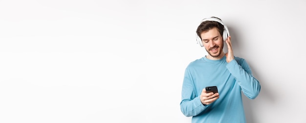 Hombre sonriente alegre escuchando música y mirando el mensaje de lectura del teléfono inteligente en el teléfono de pie en