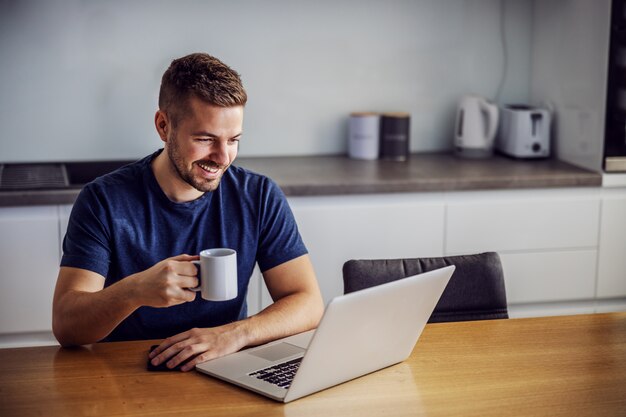 Hombre sonriente alegre joven sentado en la mesa dinging, sosteniendo la taza con café recién hecho por la mañana y mirando la computadora portátil. Está recibiendo me gusta por publicar en las redes sociales.