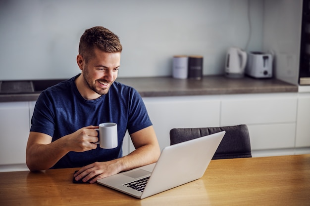 Foto hombre sonriente alegre joven sentado en la mesa dinging, sosteniendo la taza con café recién hecho por la mañana y mirando la computadora portátil. está recibiendo me gusta por publicar en las redes sociales.