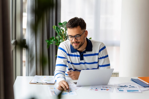 Hombre sonriente con anteojos sentado en el escritorio en la oficina mirando datos mientras trabaja en su computadora portátil