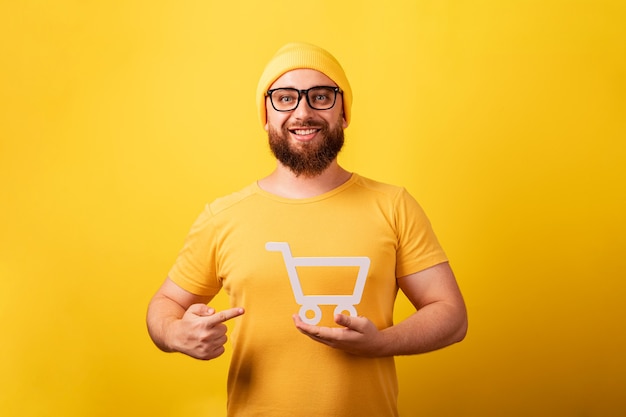 Hombre sonriente apuntando al símbolo del carrito de la compra en la mano sobre fondo amarillo, viernes negro y concepto de grandes ventas