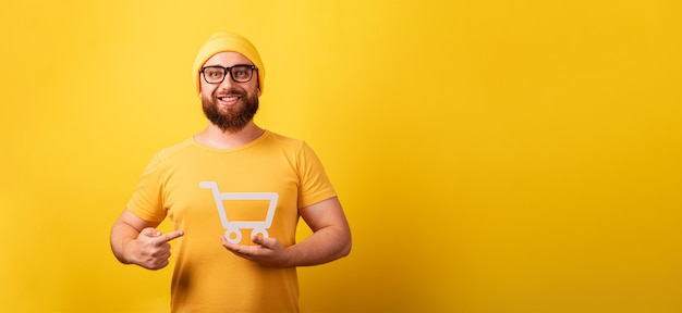 Hombre sonriente apuntando al símbolo del carro de la compra en la mano sobre fondo amarillo, viernes negro y concepto de grandes ventas, diseño panorámico