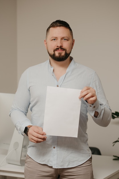 Un hombre sonriente con barba está de pie con una hoja vacía de papel blanco cerca de una computadora de escritorio de acero en casa