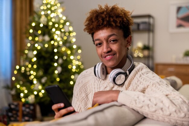 Un hombre sonriente con cabello afro claro rizado se sienta en un sofá con auriculares inalámbricos y teléfono