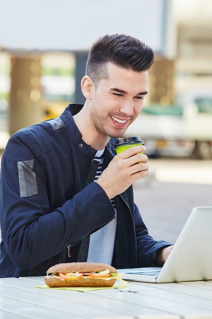 Hombre sonriente con café y sandwich trabajando en la computadora portátil